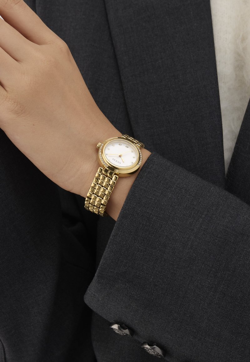 Frauenhandgelenk mit einer goldfarbenen Guess-Uhr mit weißem Zifferblatt und Kristallmarkierungen, bekleidet mit dem Ärmel eines dunkelgrauen Blazers.