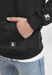 Main dans la poche d'un sweat à capuche Starter Black Label noir, jean bleu clair, manche du sweat avec logo étoile blanche brodée visible.