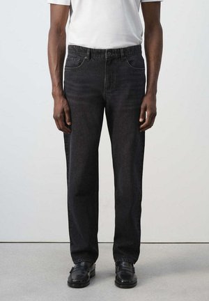Hombre con vaqueros negros de corte recto, zapatos de cuero negros y una camisa blanca de manga corta, de pie frente a un fondo liso y claro.