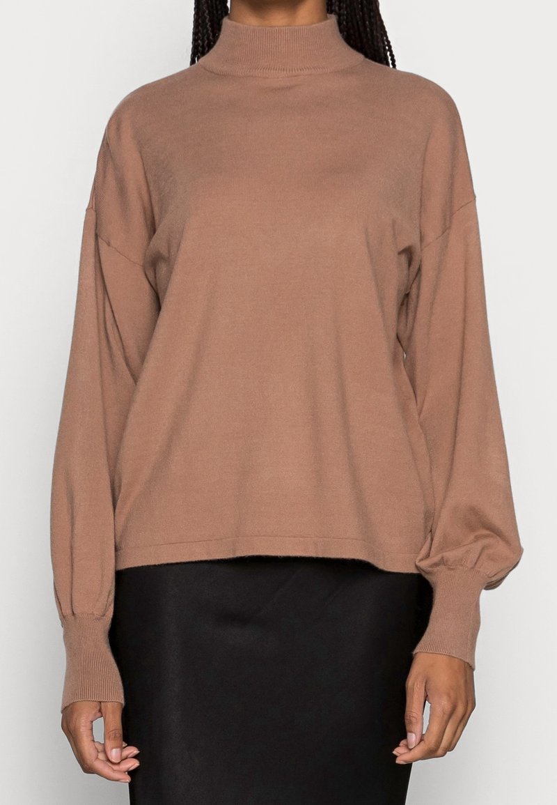 Pull marron à manches longues avec col montant et manches retroussées porté par une personne aux cheveux tressés, associé à une jupe noire.