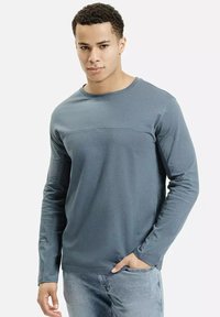 Młody mężczyzna w dopasowanej, niebieskiej koszuli z d ługim rękawem i jasnoniebieskich dżinsach, stojący z jedną ręką w kieszeni na jednolitym tle.