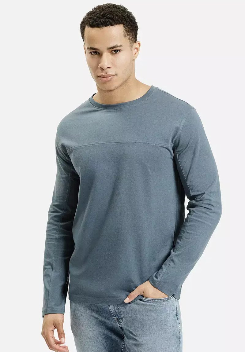 Młody mężczyzna w dopasowanej, niebieskiej koszuli z długim rękawem i jasnoniebieskich dżinsach, stojący z jedną ręką w kieszeni na jednolitym tle.