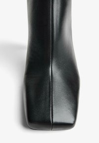 Botină de piele neagră până la gleznă, cu vârf pătrat, textură netedă, cusături proeminente și un siluetă elegantă.