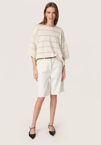 Gehaakte crème oversized trui met roze strepen boven witte knielange shorts en zwarte puntige hakken. Eenvoudig ontwerp, lichtgewicht textuur.