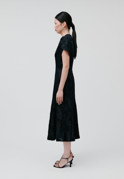 Schwarzes blütenmuster Lace-Kleid mit kurzen Ärmeln, schwingendem Saum und subtiler Textur, kombiniert mit schwarzen Absatzsandalen mit dünnen Riemen.