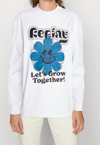 Langärmliges weißes Baumwoll-T-Shirt mit einem blauen Blumenmotiv und schwarzem Text, der in retro Schriftart "Lass uns gemeinsam wachsen!" sagt.