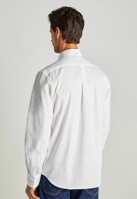 Camicia bianca a maniche lunghe con collettobutton-down, dettaglio con piega sul retro e orlo arrotondato, realizzata in tessuto morbido, indossata con jeans blu.