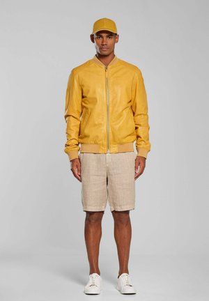 Mężczyzna stojący, ubrany w żółtą kurtkę i czapkę, beżowe szorty i białe trampki, na jednolitym szarym tle.