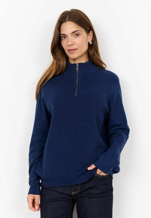 Mörkblå stickad tröja med hög krage och halv dragkedja, med ribbad textur och långa ärmar, kombinerad med mörka jeans.