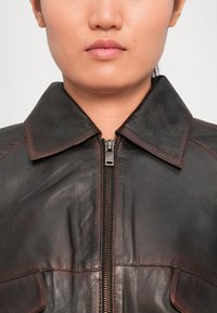 Közeli kép egy sötétbarna bőrdzsekit viselő személyről, gallérral és középső fém cipzárral, egy sima háttér előtt.