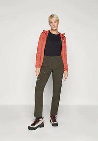 Κόκκινο μπουφάν με φερμουάρ, μαύρη μπλούζα και ελαιοπράσινα παντελόνια με τσέπες. Πεζοπορικά μποτάκια με κόκκινες κορδόνες. Οριζόντιο φόντο, πόζα όρθια.