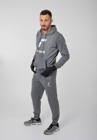 Grå hoodie med en frontlomme og logo, parret med matchende sweatpants og sorte handsker. Modellen bærer grå sneakers.