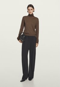 Donna con un maglione a collo alto marrone, pantaloni neri a gamba larga, scarpe nere, che tiene una piccola borsa nero, in piedi su uno sfondo neutro.