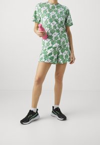 Henkilö, joka on pukeutunut vihreälehtiseen yhteensopivaan paitaan ja shortseihin, pitää pinkkiä vesipulloa, seisoo mustissa lenkkarissa, joissa on valkoiset pohjat.