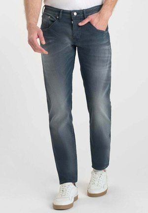 Jeans pour hommes en denim bleu foncé, coupe slim, texture usée, cinq poches, fermeture à bouton en métal, associés à des baskets blanches.