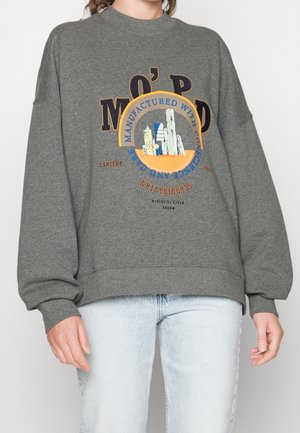 Personne portant un sweat-shirt gris avec un graphisme de paysage urbain et du texte, associé à un jean bleu clair, debout devant un fond uni.