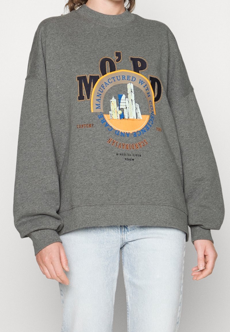 Personne portant un sweat-shirt gris avec un graphisme de paysage urbain et du texte, associé à un jean bleu clair, debout devant un fond uni.