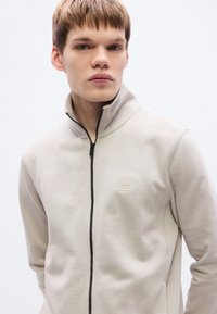 Μπεζ ζακέτα με φερμουάρ κατασκευασμένη από απαλό ύφασμα, με ψηλό γιακά και μικρό τετράγωνο λογότυπο στο αριστερό στήθος. Μαύρη λεπτομέρεια φερμουάρ.