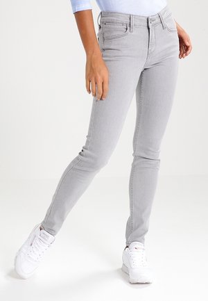 Persoon die lichtgrijze skinny jeans, witte sneakers en een lichtblauw topje draagt, staand met één been naar voren gestrekt.