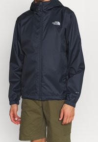 Mann trägt eine marineblaue North Face Jacke mit Reißverschluss und elastischen Bündchen sowie olivgrüne Cargo-Shorts vor einem schlichten Hintergrund.