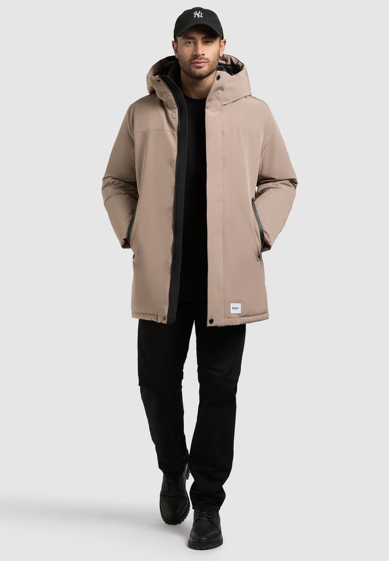 Parka beige con capucha, cierre oculto, bolsillos laterales y acentos negros. Llevada sobre una camiseta negra y combinada con pantalones y botas negras.
