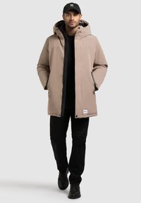 Parka beige con cappuccio, dotata di zip nascosta, tasche laterali e dettagli neri. Indossata sopra una maglietta nera e abbinata a pantaloni neri e stivali.