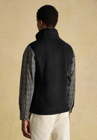 Melnas vilnas veste ar augstu apkakli, ar uzrakstītu zīmola nosaukumu, vilkta virs raibā krekla, izlikta uz neitrāla fona.