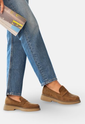 Personne portant un jean bleu et des mocassins en daim marron, tenant un magazine plié dans une main, debout devant un fond uni.