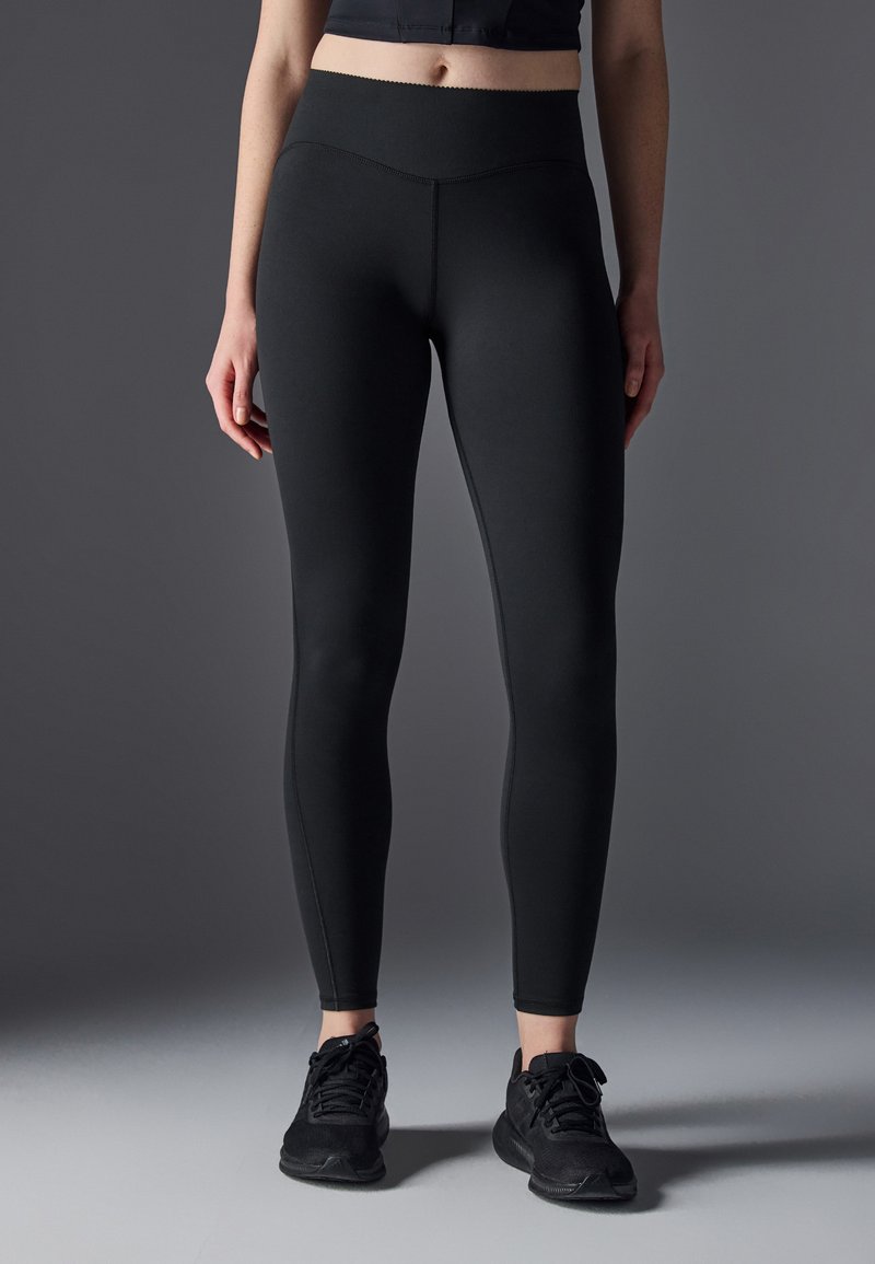 Leggings noirs à taille haute avec une texture lisse et extensible. Les caractéristiques incluent un design ajusté et des coutures épurées, associés à des chaussures de sport noires.