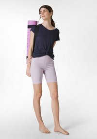 Marinblå knuten T-shirt kombinerad med lila cykelshorts. En rullad yogamatta med axelrem vilar mot modellens axel.