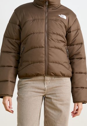 Doudoune marron avec fermeture éclair frontale, poches latérales zippées, et logo The North Face sur la poitrine, portée avec un pantalon beige à rayures blanches.