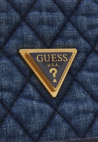 Sac en denim avec texture matelassée, arborant un emblème en triangle doré avec "GUESS" et un point d'interrogation. Couleur bleu profond avec un motif subtil.