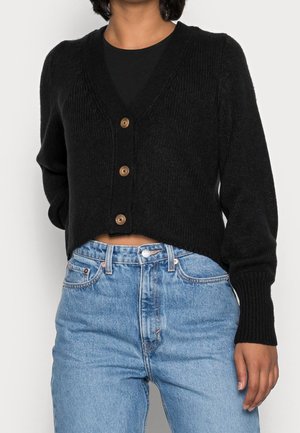 Femme portant un cardigan noir court boutonné par-dessus un haut noir et un jean taille haute bleu clair, debout devant un fond uni.