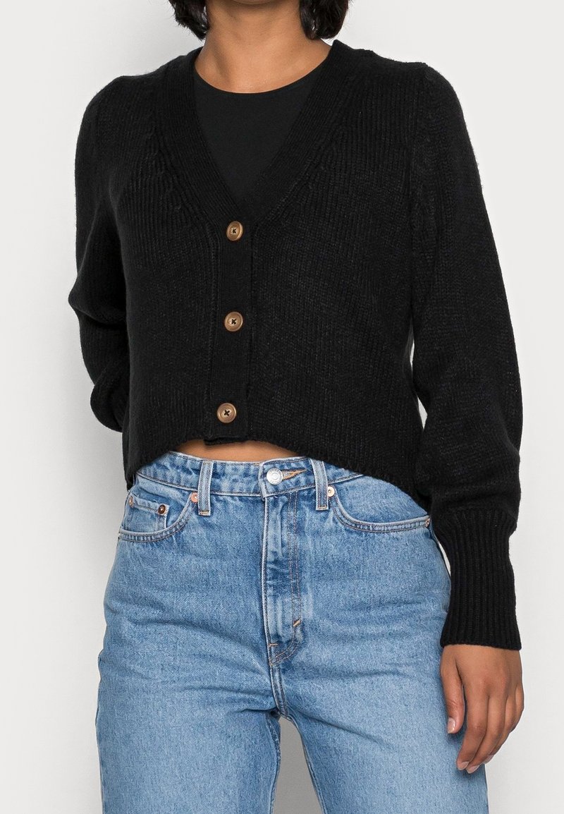 Femme portant un cardigan noir court boutonné par-dessus un haut noir et un jean taille haute bleu clair, debout devant un fond uni.