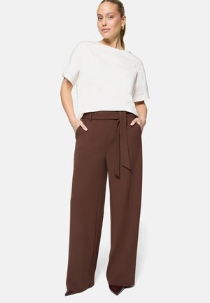 Femme portant un chemisier blanc à manches courtes et un pantalon large marron taille haute avec ceinture, debout les mains dans les poches.