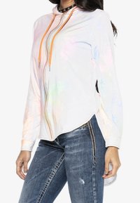 Haut zipé blanc avec des cordons orange, présentant une texture brillante et iridescente et des manches longues, associé à un jean bleu déchiré.
