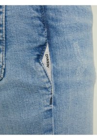Pantalones vaqueros de mezclilla azul claro con textura deslavada. Presentan una etiqueta visible con la palabra "CHASIN'" en la costura lateral. Diseño clásico de cinco bolsillos.