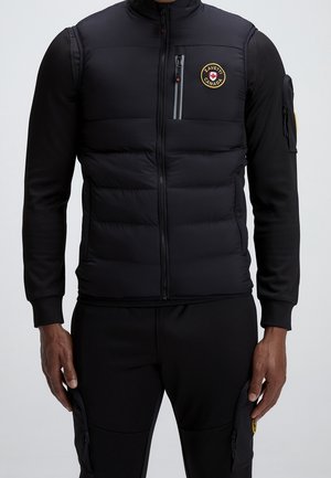 Homme portant une veste matelassée noire avec fermeture éclair frontale et un patch rond avec le logo Zavetti Canada, sur une chemise noire à manches longues et un pantalon noir avec poches zippées.