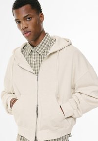 Beige Kapuzenpullover mit Reißverschluss in lässiger Passform, ausgestattet mit einer Kängurutasche und einer Kapuze mit Kordelzug, über einem grün-karierten Hemd getragen.