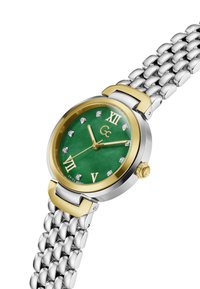 Ceas argintiu și auriu, cu un cadran verde, cifre romane și accente din diamant. Dispune de o brățară din plasă de oțel inoxidabil.