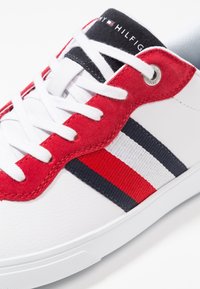 Baskets blanches avec des accents en daim rouge, lacets blancs, et détail en tissu rayé bleu marine, blanc et rouge, marquées du logo Tommy Hilfiger sur la languette.