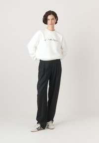 Fehér pulóver "Lemonade" felirattal, fekete, redőzött nadrággal és fehér, fekete csíkos sportcipővel párosítva. Egyszerű háttér.