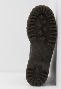 Semelle d'une botte noire avec un motif de bande de roulement robuste, montrant des crampons profonds et des grips en forme de croix sur une surface blanche.