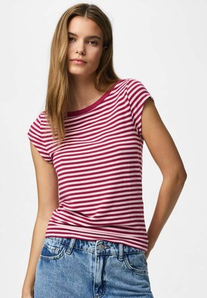 Ung kvinne med en rødhvit stripet kortermet skjorte og blå denimjeans, står foran en ensfarget hvit bakgrunn.