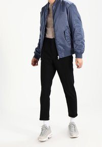 Hellblaue Bomberjacke mit glattem Finish, graues Hemd, schwarze, zulaufende Hose und graue Sneaker mit klobiger Sohle. Weiße Socken sichtbar.