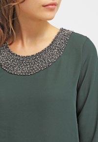 Haut vert foncé avec un large col orné de perles présentant des motifs complexes en argent et noir. Texture de tissu lisse ; manches longues.