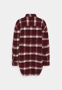 Chemise en flanelle à carreaux rouge et blanc, avec des manches longues, un col et une coupe décontractée. Le matériau semble doux et texturé.