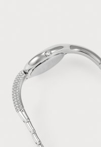 Silberne Uhr mit einem geschwungenen Gehäuse und einem zentralen Kristallakzent. Armband verziert mit kleinen, runden, funkelnden Details. Glatte, polierte Oberfläche.