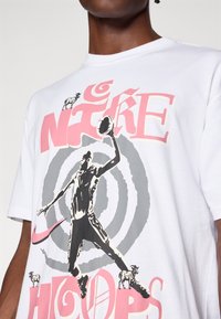 Witte katoenen t-shirt met een zwarte afbeelding van een basketballer, vergezeld van roze en grijze tekst met daarop "Nike Hoops" en kleine illustraties van een geit.