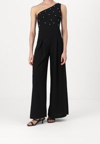 Zwarte jumpsuit met een asymmetrisch ontwerp met één schouder, versierd met witte parels, en brede pijpen. Gladde textuur.